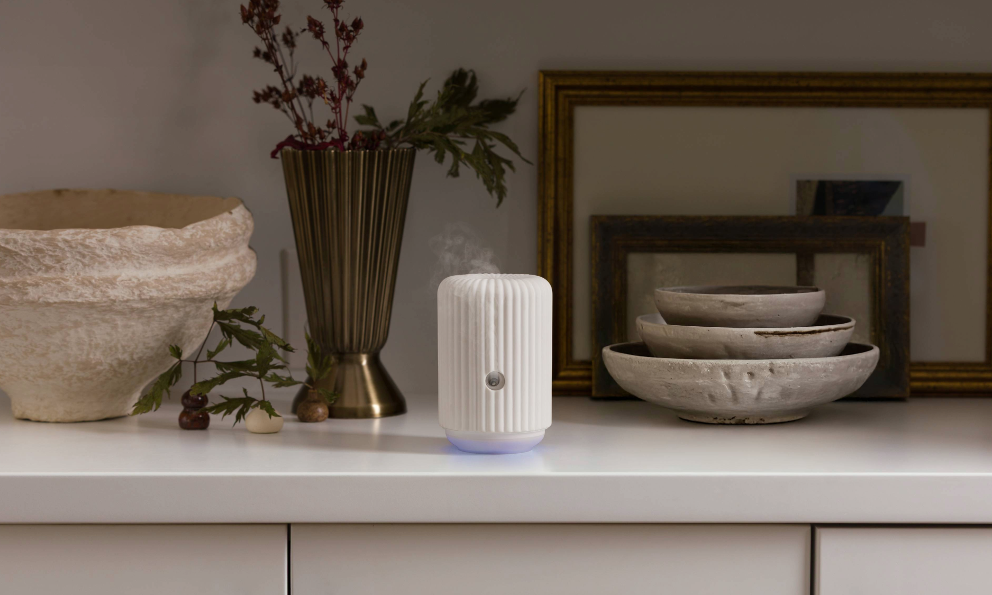 Smart Diffuser in white with a ribbed ceramic design, softly glowing at the base with visible mist rising, styled on a neutral tabletop beside decorative bowls and brass accents.