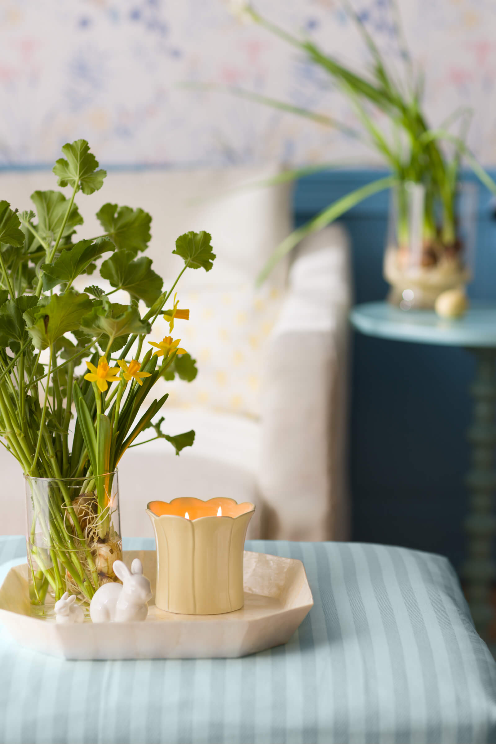 A lit Wild Honeysuckle & Willow Scented Candle sits in a tray on a table beside a rabbit figurine and a vase of honeysuckles and greenery.