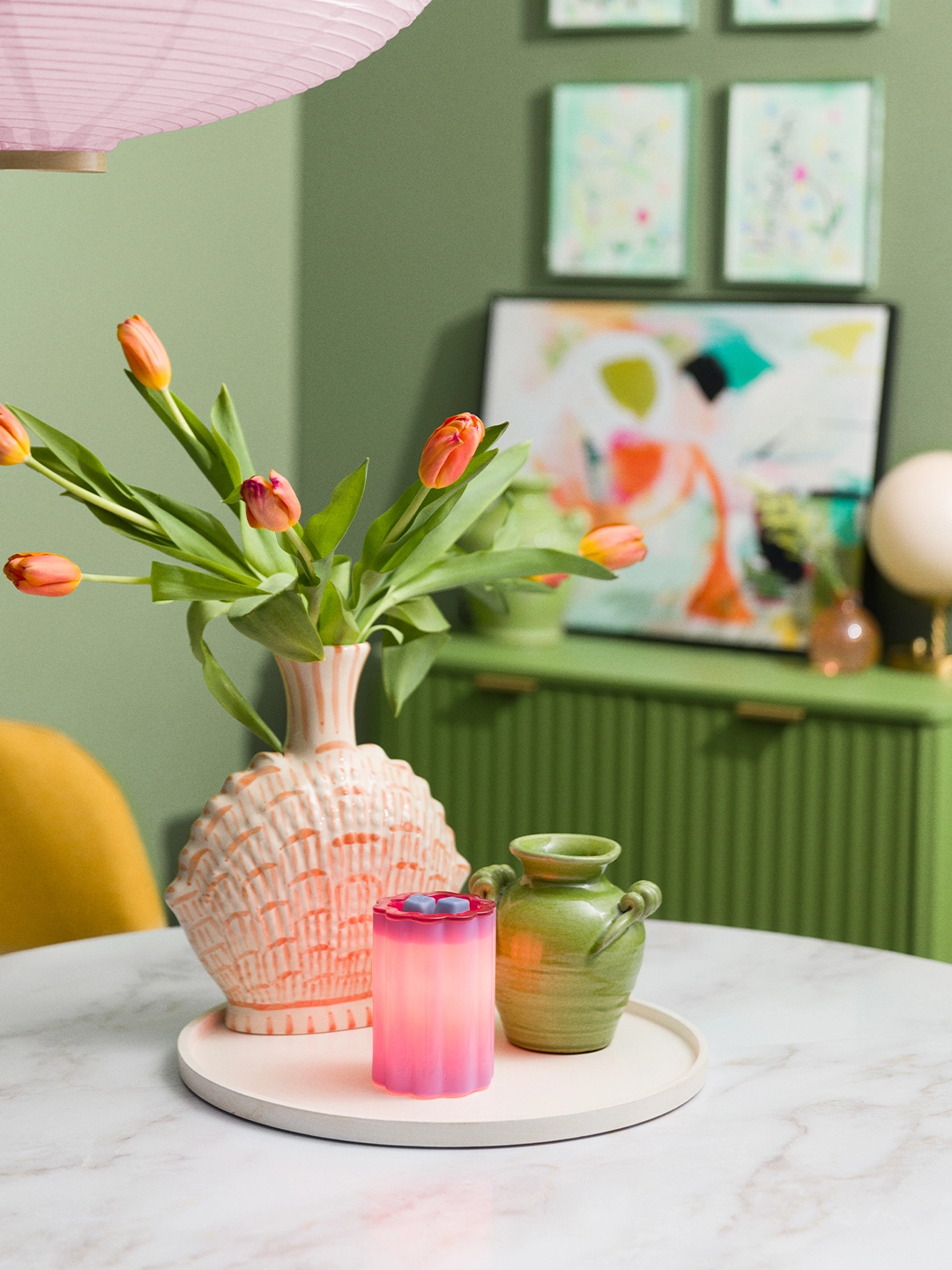 Bloomie-Purple glass warmer lit to reveal a soft pink-to-purple ombré glow, styled on a round tray beside decorative vases and tulips.