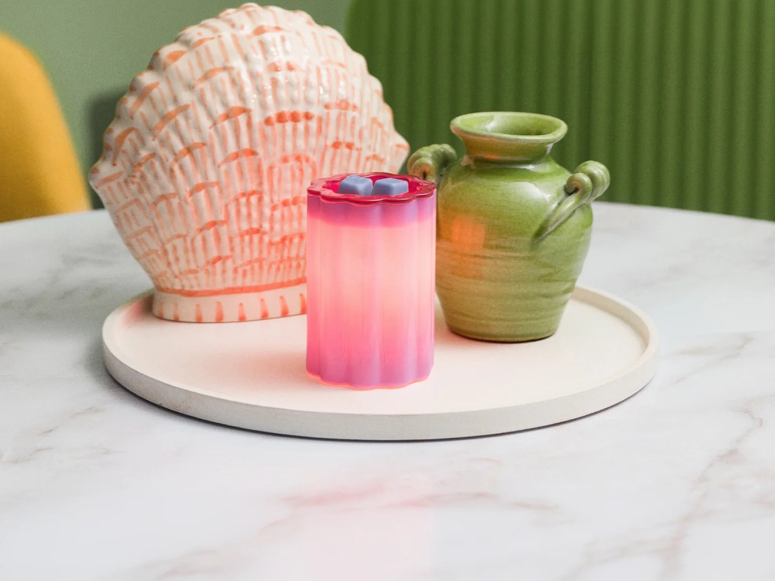A Petal – Purple Warmer is displayed on a white tray with brightly coloured vases, tulips and a green cabinet in the background. 