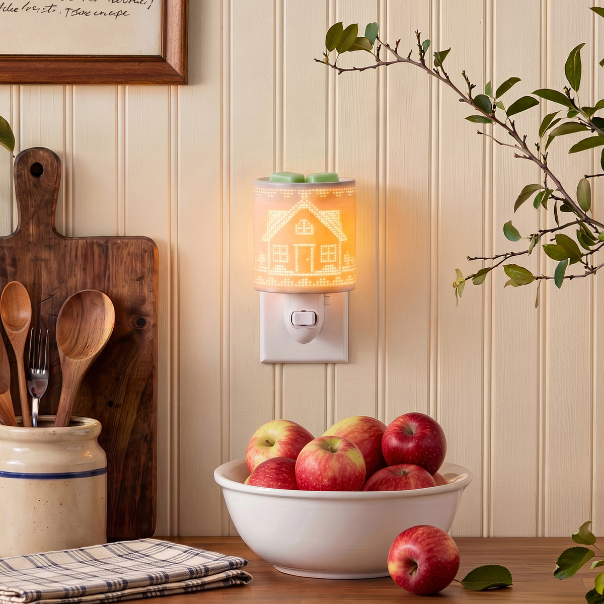 Minicalentador Our House iluminado con un acogedor diseño de casita, conectado a una pared de cocina de madera blanca sobre un tazón de manzanas rojas, con cucharas de madera en un tarro, una tabla para picar y un trapo a cuadros