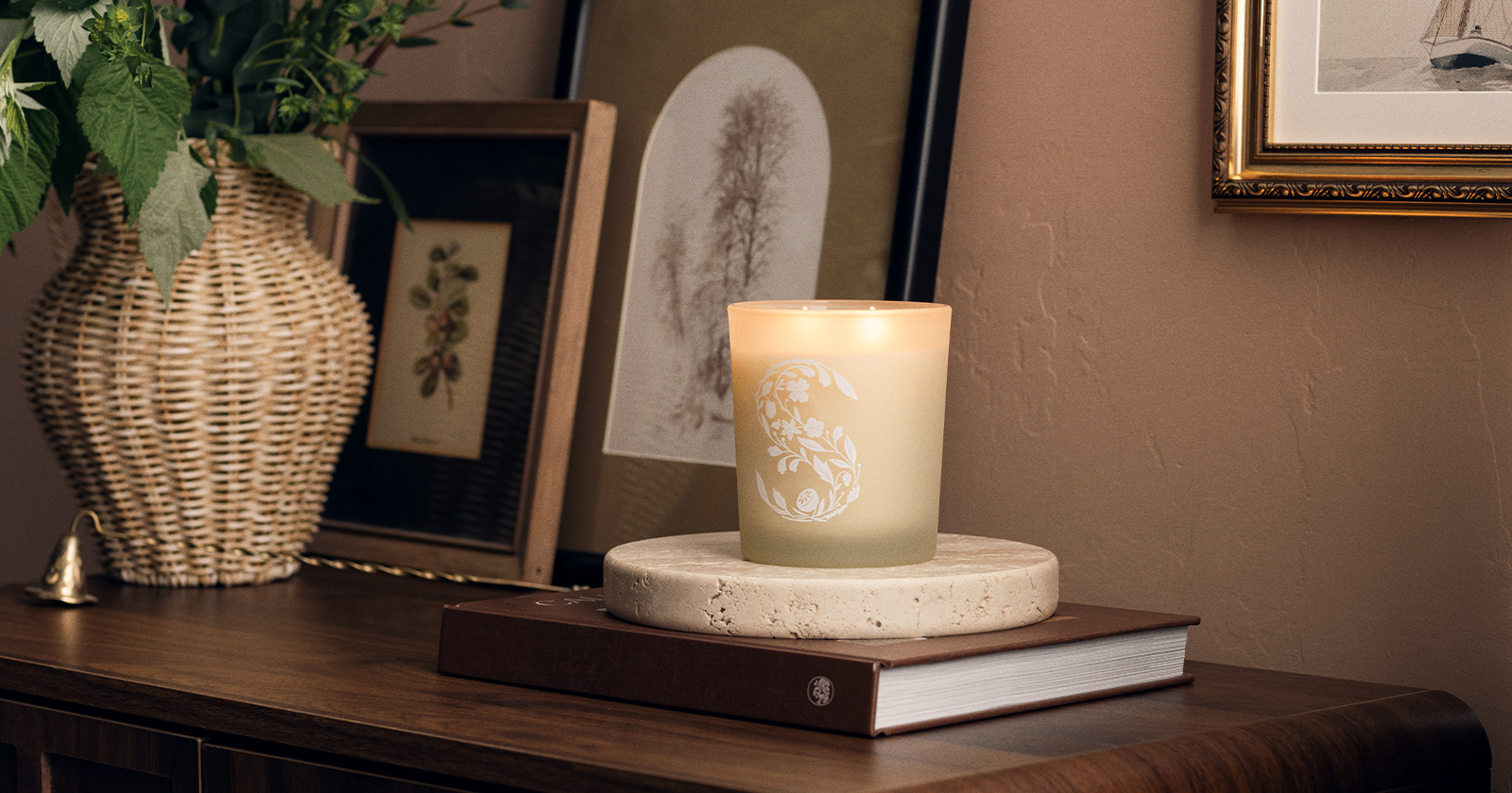 Scentsy frosted glass candle with white floral S logo illuminated on round stone coaster, displayed on stacked books with botanical art prints and wicker vase