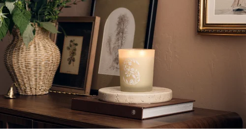 Scentsy scented candle displayed on a wooden surface beside decorative vases and framed artwork in a warm, neutral home setting.