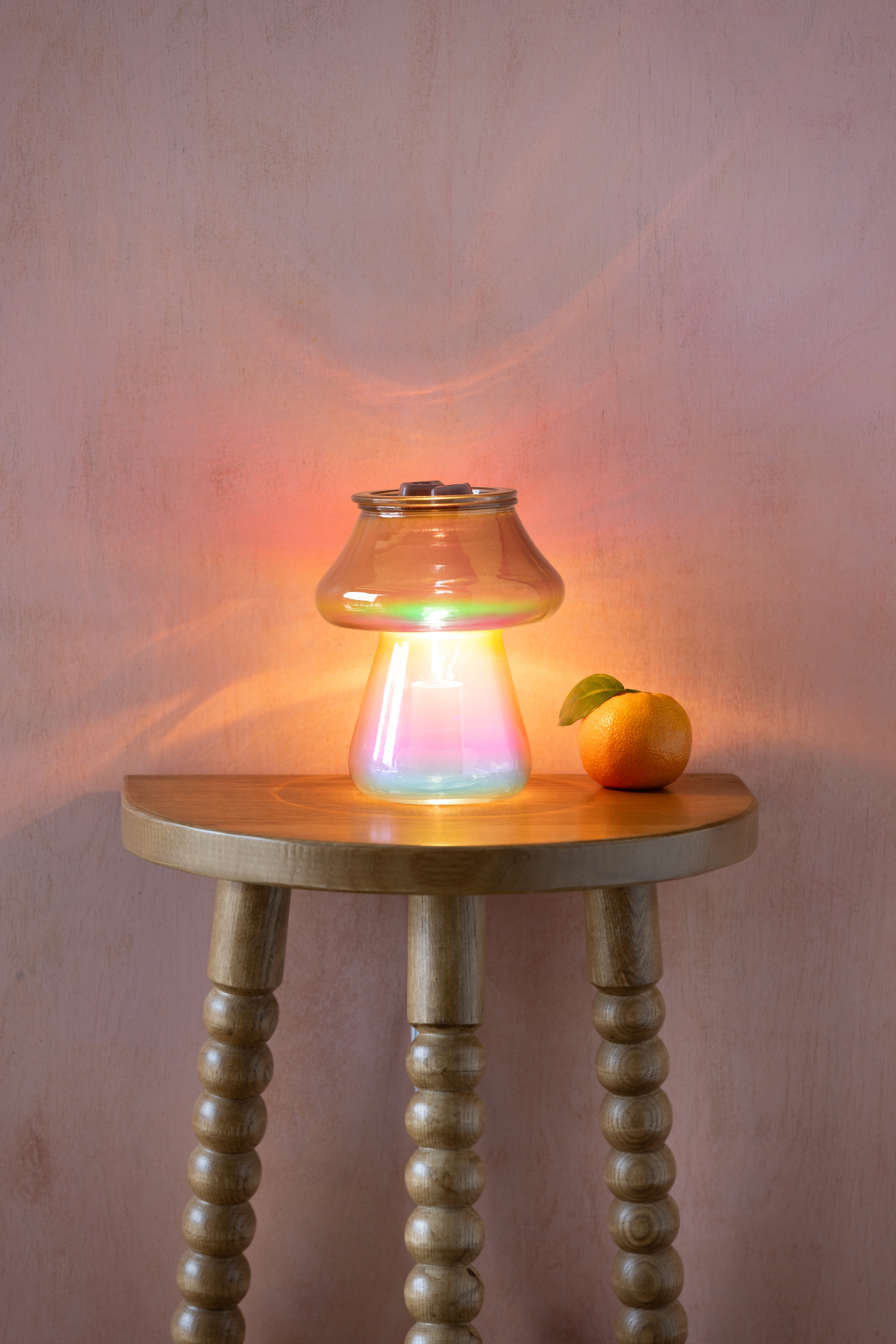 Amanita Warmer by Scentsy on a wooden side table, its bronze-ombre glass mushroom design glowing softly and surrounded by warm ambient light, with a fresh mandarin placed beside it.
