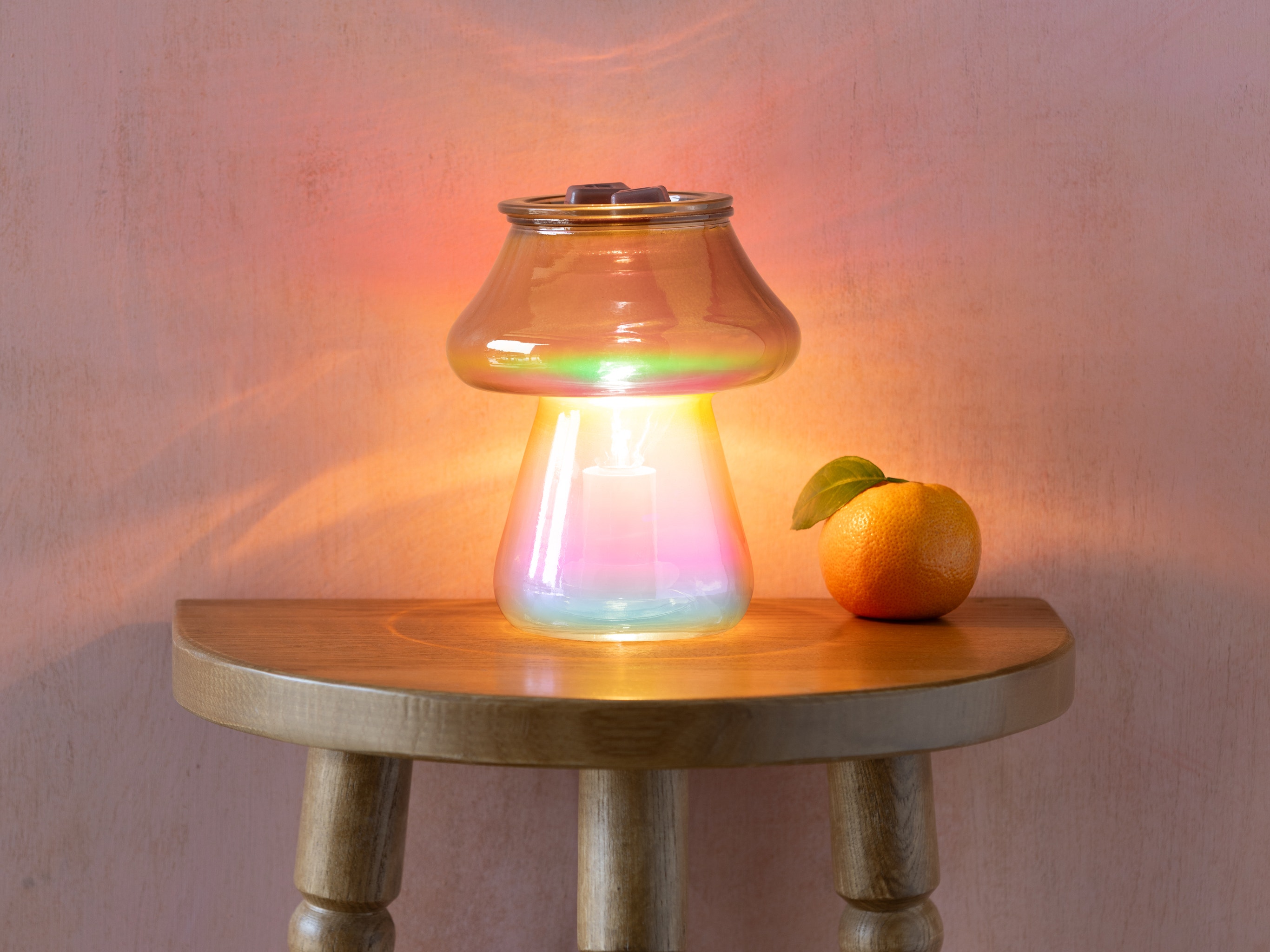 Amanita Warmer by Scentsy on a wooden side table, its bronze-ombre glass mushroom design glowing softly and surrounded by warm ambient light, with a fresh mandarin placed beside it.