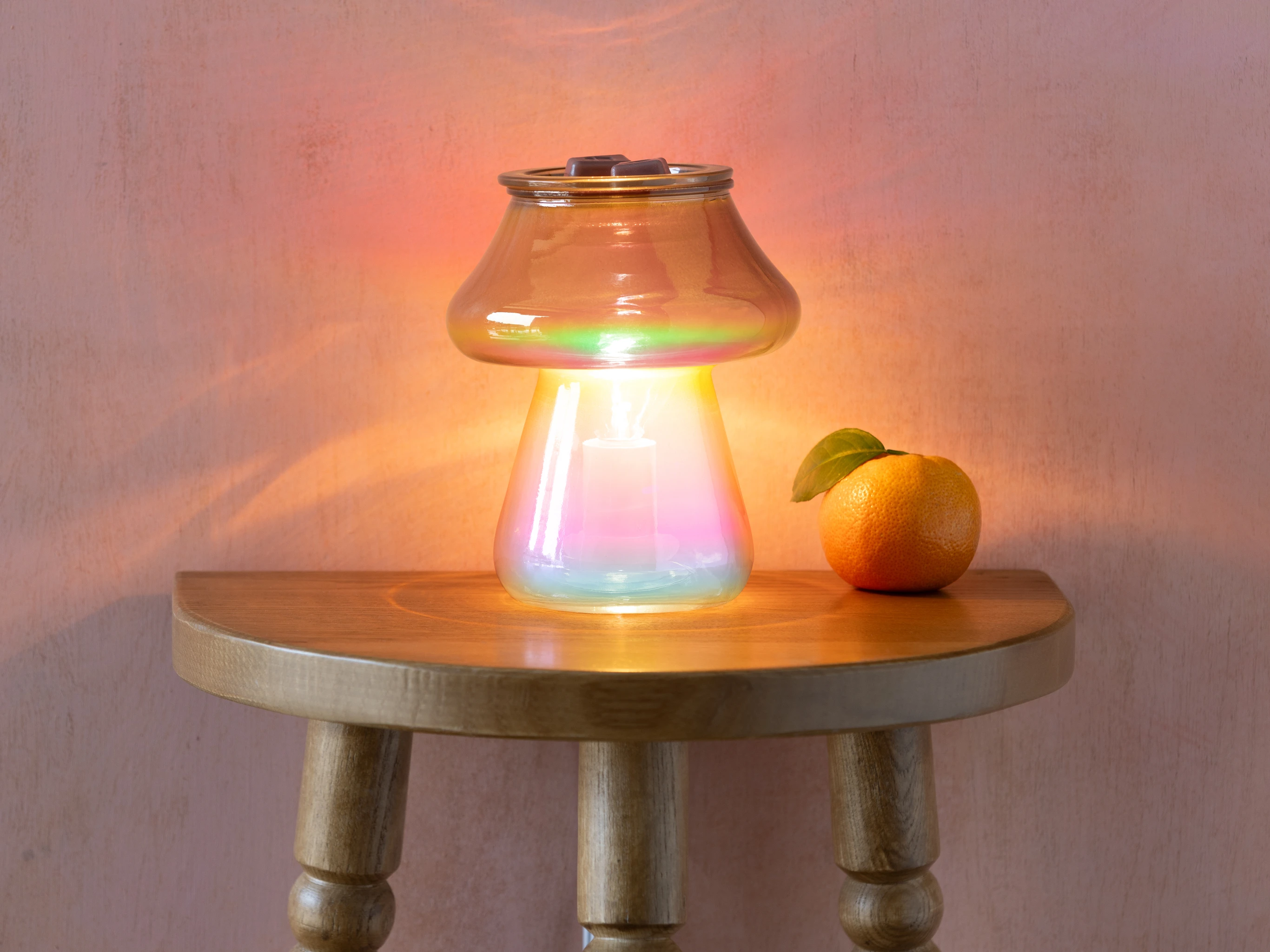 Amanita Warmer by Scentsy on a wooden side table, its bronze-ombre glass mushroom design glowing softly and surrounded by warm ambient light, with a fresh mandarin placed beside it.