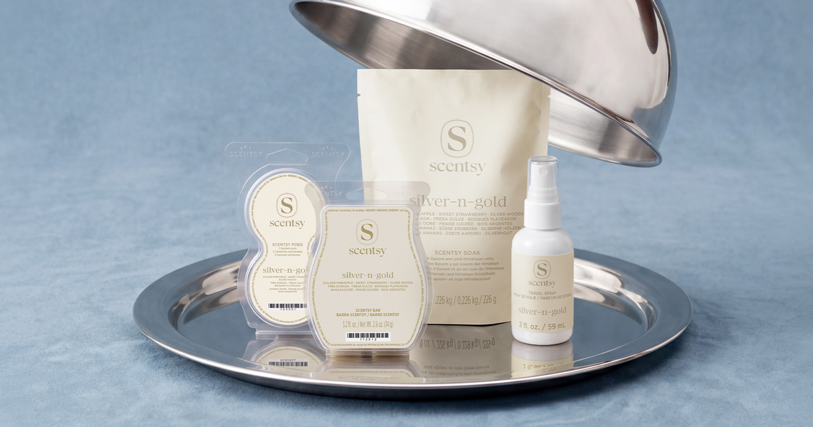 A hand lifts the cover from a silver cloche tray containing products in the Silver-N-Gold fragrance. 