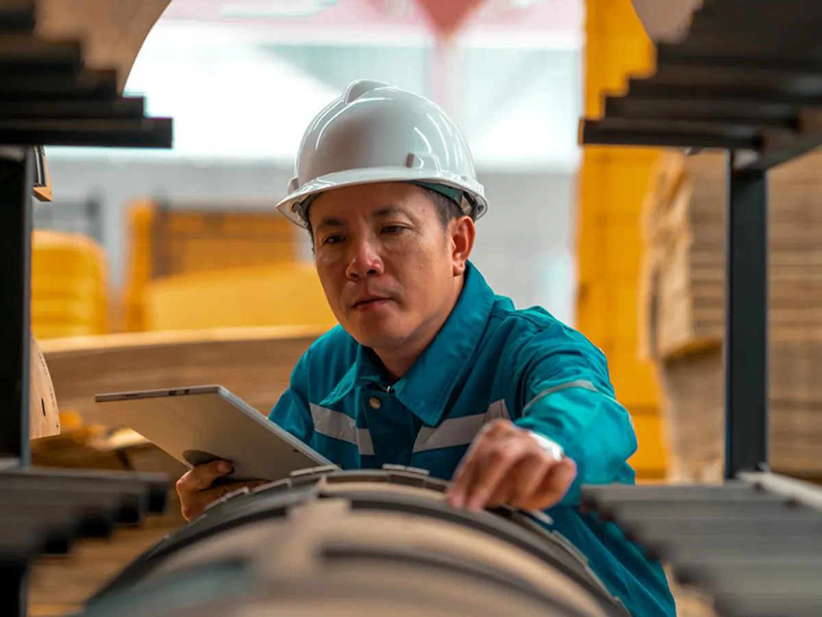 Worker in a hard hat and reflective jacket inspecting an industrial conveyor belt with a tablet in hand.