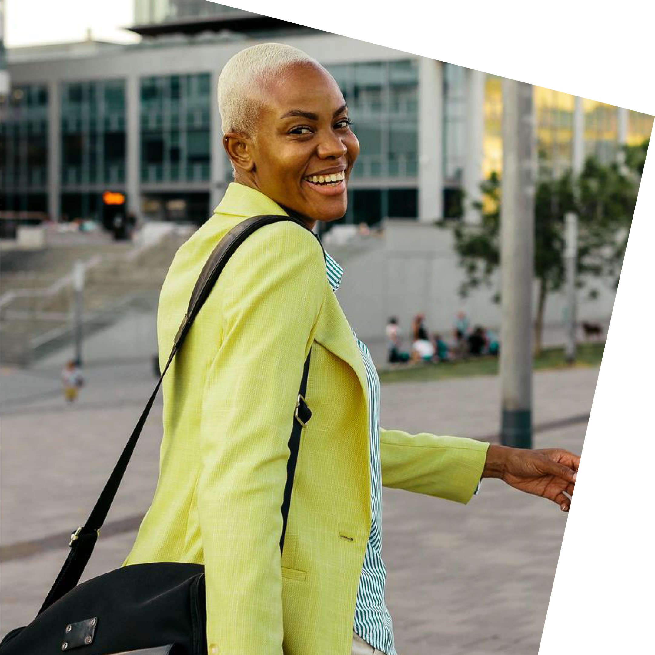 Smiling person with short hair in a yellow jacket and striped shirt, carrying a black bag. Urban background with modern buildings, stairs, and greenery.