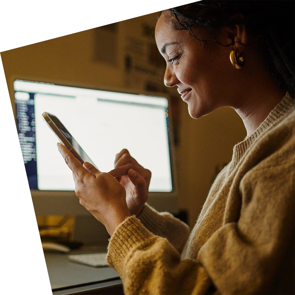 A woman in a cozy sweater uses a smartphone, smiling while sitting at a desk. A computer screen glows in the dimly lit room, creating a warm, focused atmosphere.