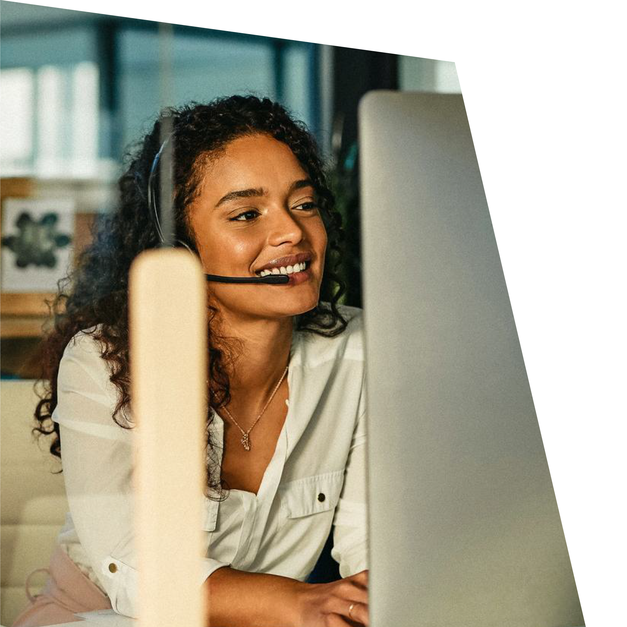 Smiling woman with curly hair wearing a headset sits at a desk, engaging with a computer screen. She appears focused and content in an office setting.