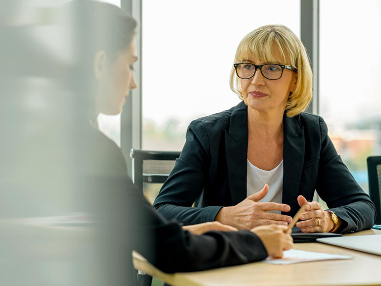 Two women in business attire having a serious discussion at a table in an office setting.