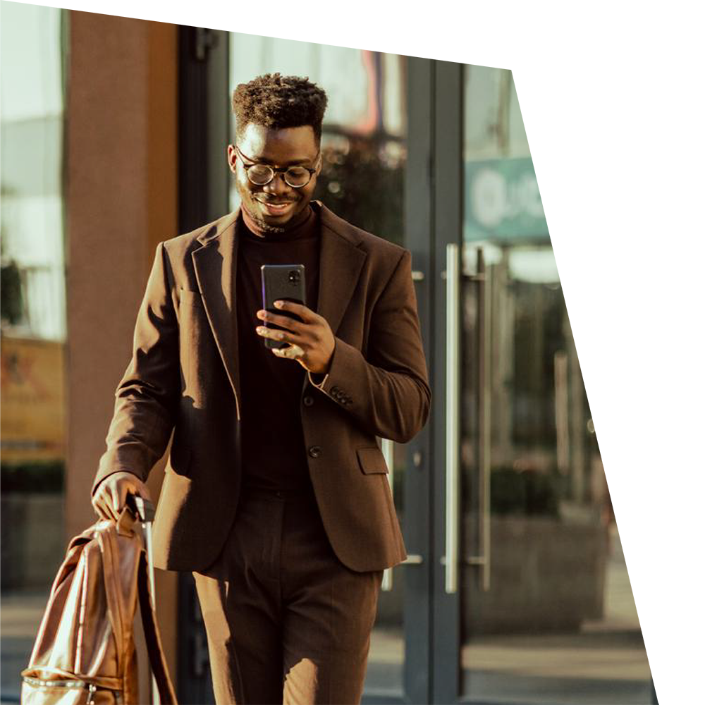 A man in a brown suit walks outside, smiling and looking at his phone. He carries a brown bag, conveying a professional and relaxed vibe in sunlight.