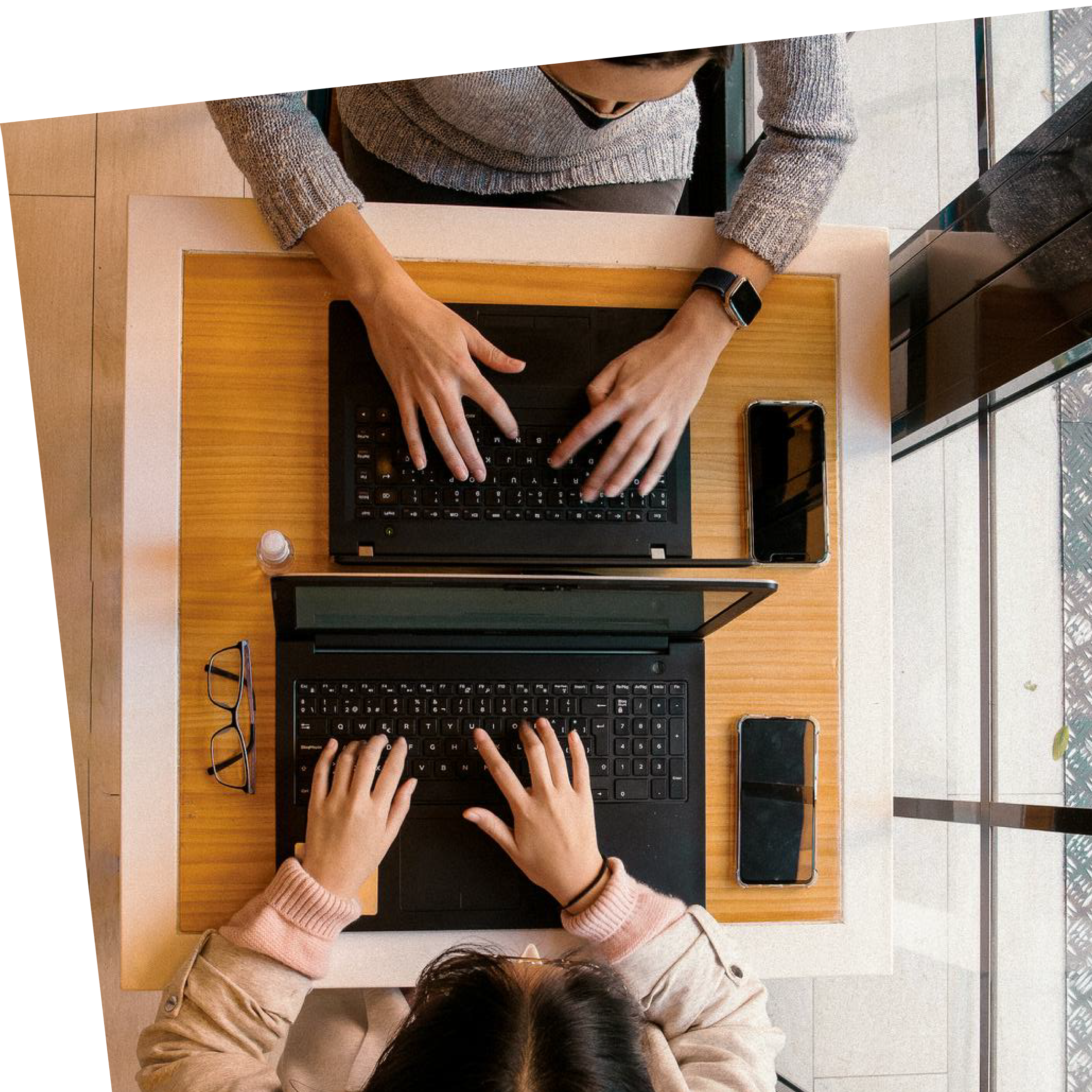 Two people typing on laptops facing each other at a wooden table. Glasses, a smartphone, and a bottle are placed nearby, suggesting focused work.