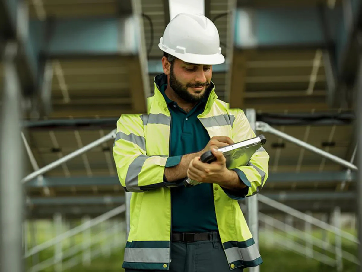 A man in a reflective jacket and hard hat uses a tablet under solar panels.