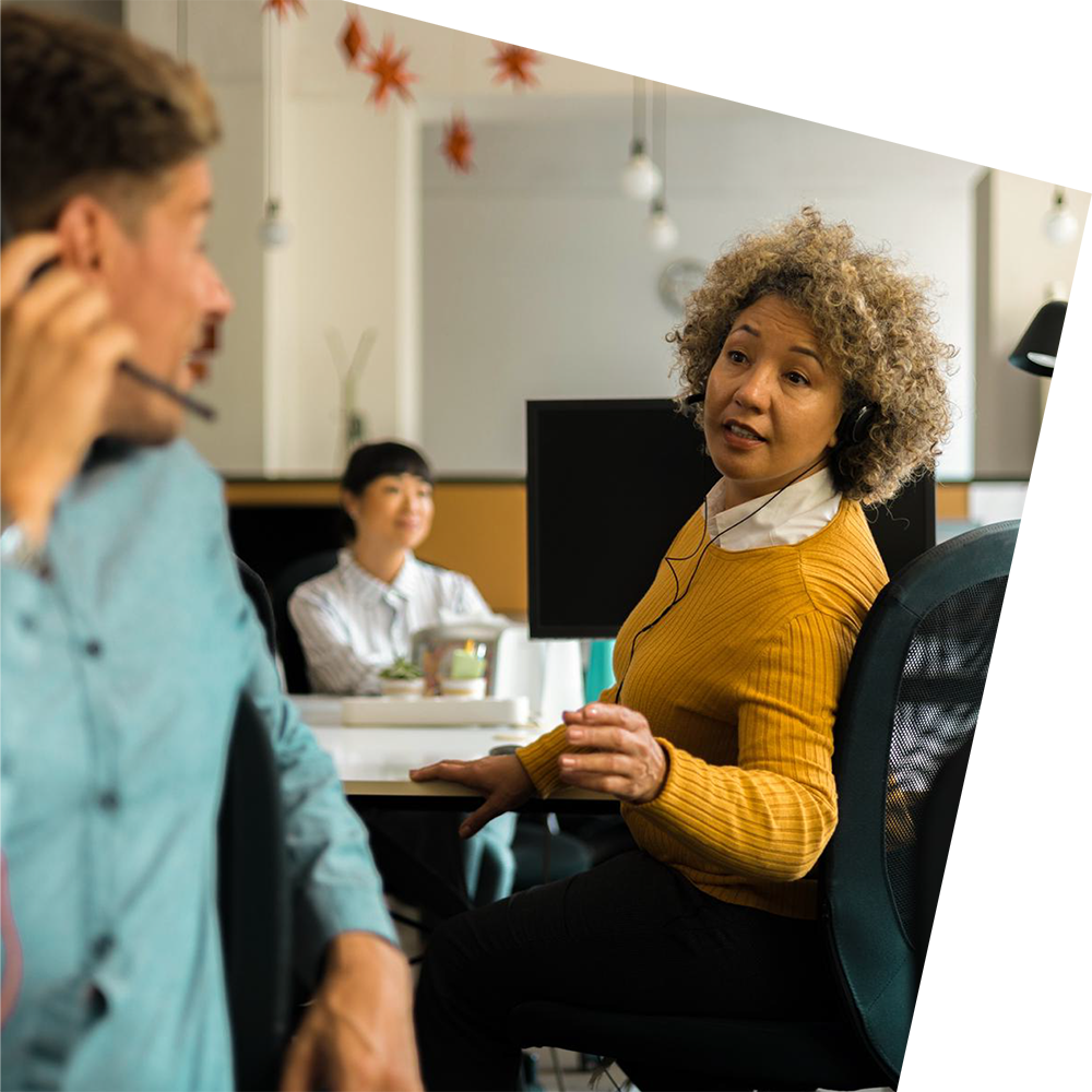 Office scene with a woman in a yellow sweater speaking to a nearby colleague wearing a headset. A third person sits in the background. Relaxed atmosphere.