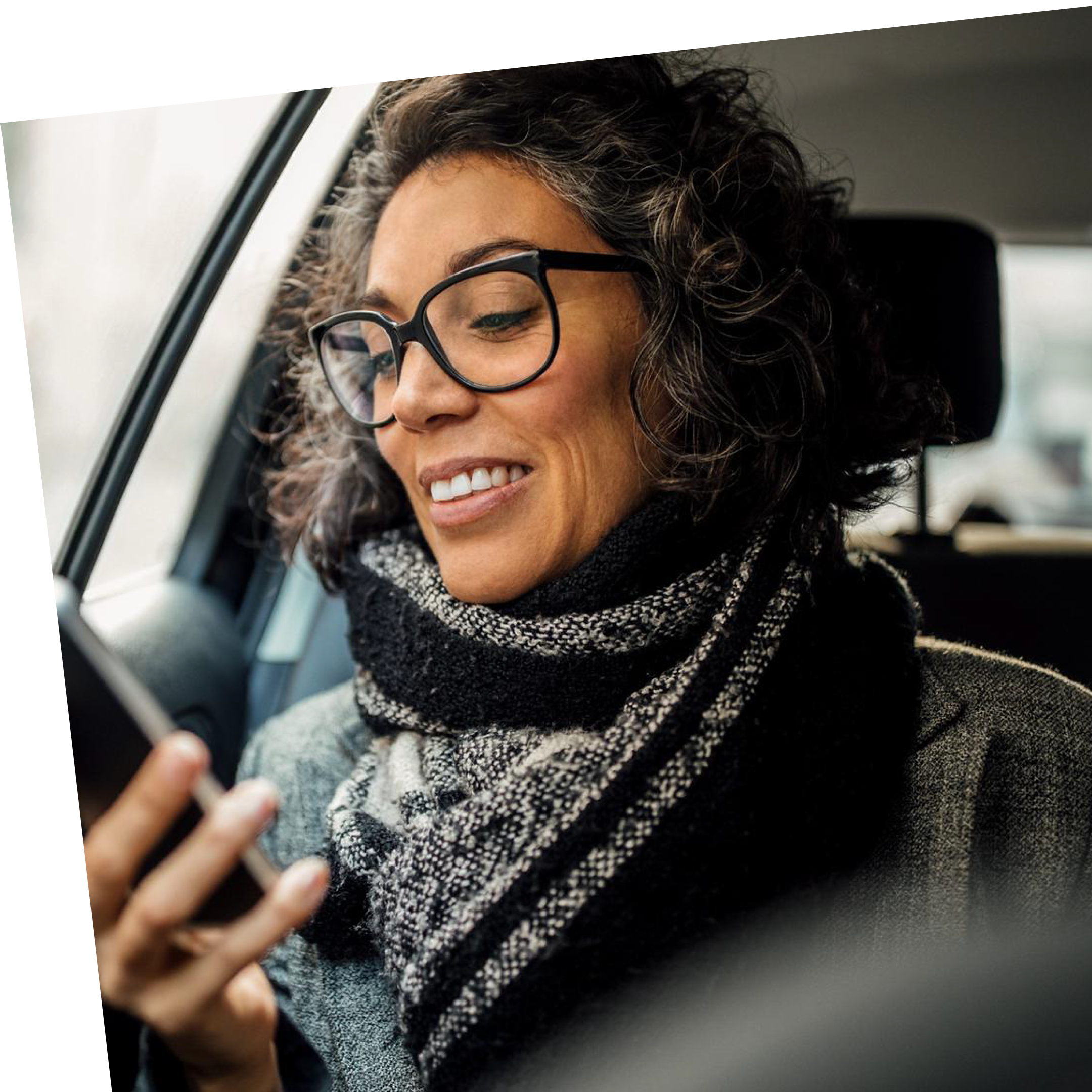 A woman with curly hair and glasses smiles while sitting in a car, wearing a cozy knit scarf, conveying a sense of warmth and contentment.