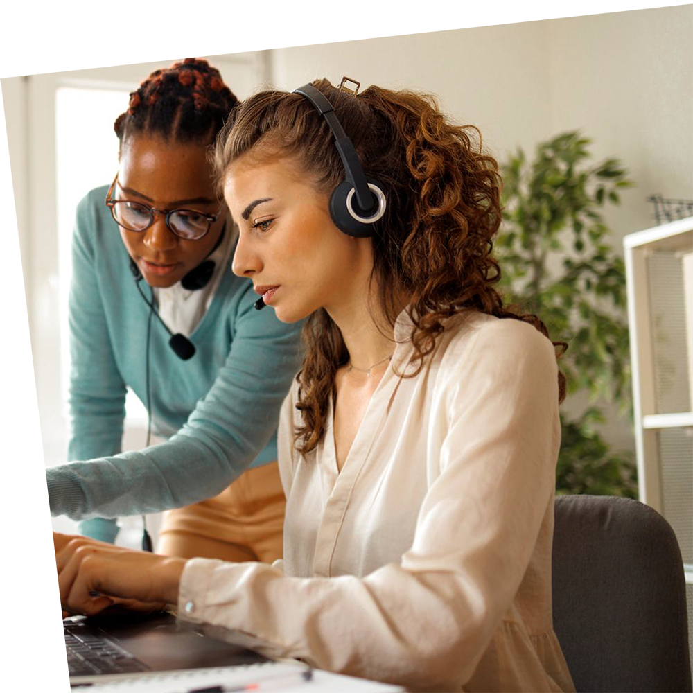 Two women at a desk, both wearing headsets. One is seated, typing on a laptop, focused, while the other stands, pointing at the screen, offering guidance. Bright, office setting.