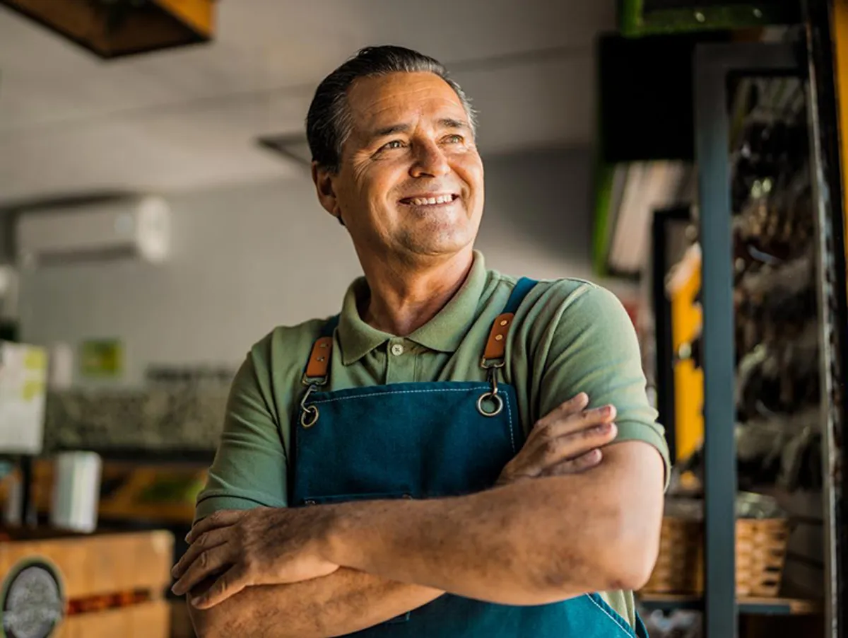 Man in a green shirt and blue apron smiles with arms crossed, standing indoors.