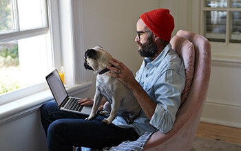 A man with a red beanie on his head sits on a chair with a laptop and pug on his lap.