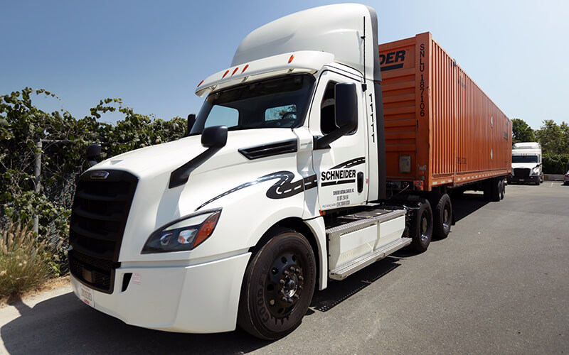 A white eCascadia semi-truck hauling an orange Intermodal container is parked at a California facility.