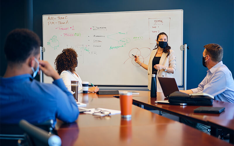A women stands at a white board while wearing a face mask and presents to a group of men and women who are also wearing face masks.