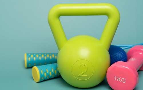 A green kettlebell, blue jump rope and set of pink hand weights arranged on the ground. 