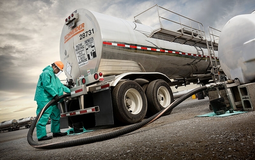 A Schneider Tanker truck driver in a blue HazMat suit unloads liquid bulk freight from a tanker truck using a large hose.