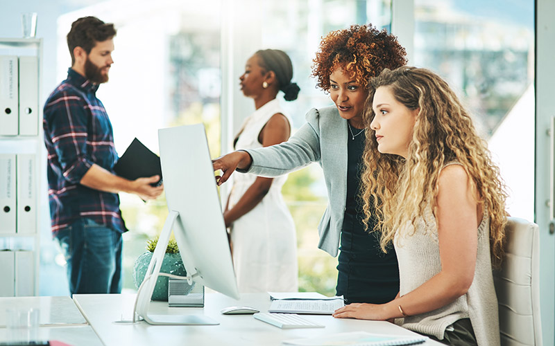 Two women standing and looking at a computer screen. One of the women is typing and the other is pointing to the screen.