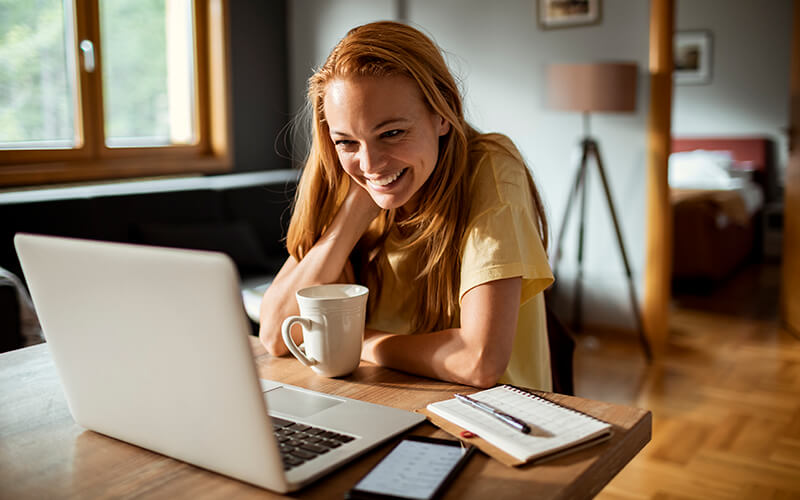 A young woman who's smiling sits at a desk with a laptop, phone, note pad and cup of coffee in front of her while working from home.