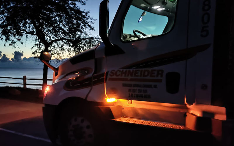 A Schneider truck with its lights on is parked at a scenic truck stop for the evening.