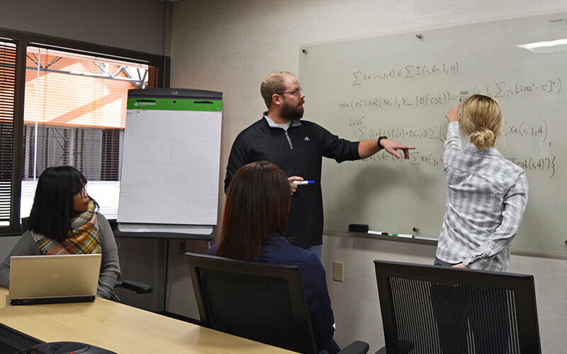 A group of Schneider associates work with equations on a glass whiteboard in a conference room.