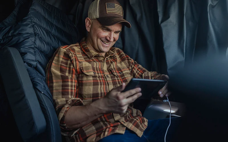 A truck driver sitting inside their semi-truck cab, using a tablet connected to a charging cable.