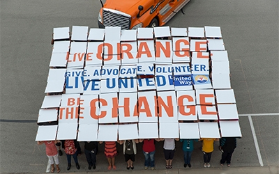 80 Schneider associates hold boards overhead that together say "Live Orange. Give. Advocate. Volunteer. Live United. Be the change."
