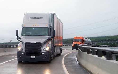 A grey Schneider Freightliner semi-truck hauling a dry van trailerdrives on an elevated highway overpass. An orange Schneider semi-truck hauling a tanker truck follows behind the first truck.