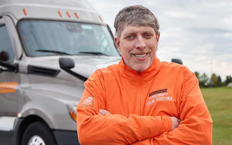 Kolin Stutcher standing outside in front of a gray Schneider semi-truck with his arms crossed in front of his chest, smiling straight ahead.