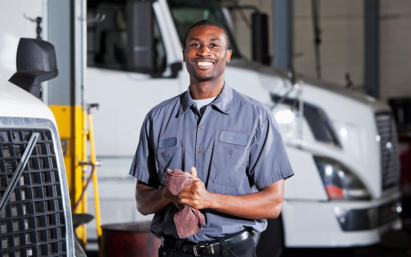 A diesel technician wipes his hands with a cloth while standing in front of two white semi-trucks inside of a shop.