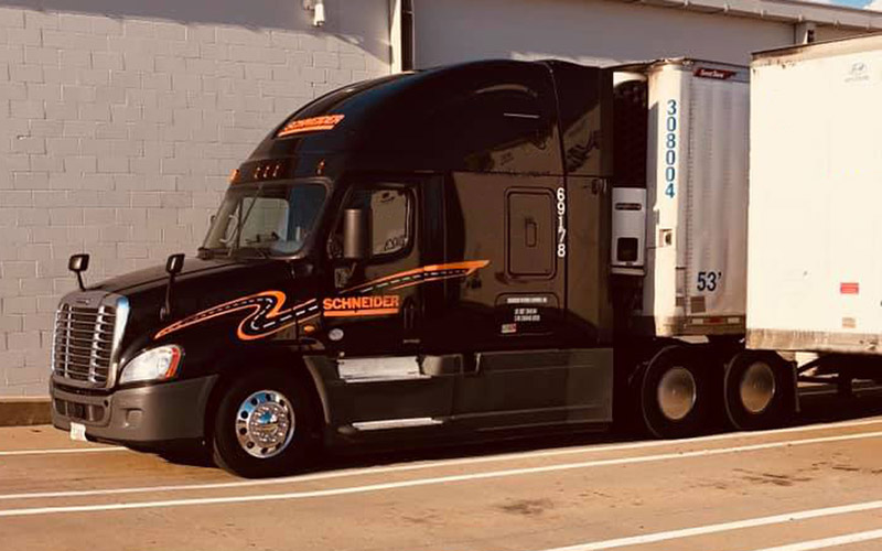 A black Schneider truck with orange and white detailing parked beside a building. The truck’s cab is attached to a white reefer trailer, which has reflective stripes and identification numbers on its side.