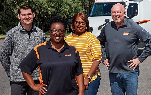 Four Schneider associates smile and pose while standing on the pavement with a Schneider semi-truck in the background