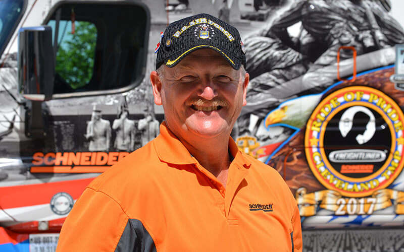 David Buck smiles wearing an orange Schneider polo and U.S. Air Force hat in front of the 2018 Ride of Pride truck