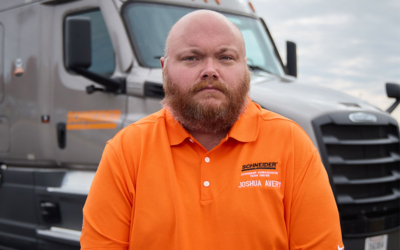 Joshua Avery standing outside in front of a gray Schneider semi-truck, looking straight ahead.