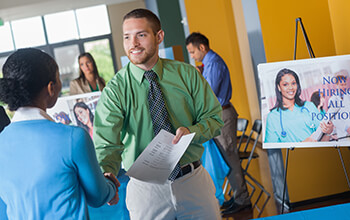 A man shakes hands with a woman and hands her his resume while attending a career fair.