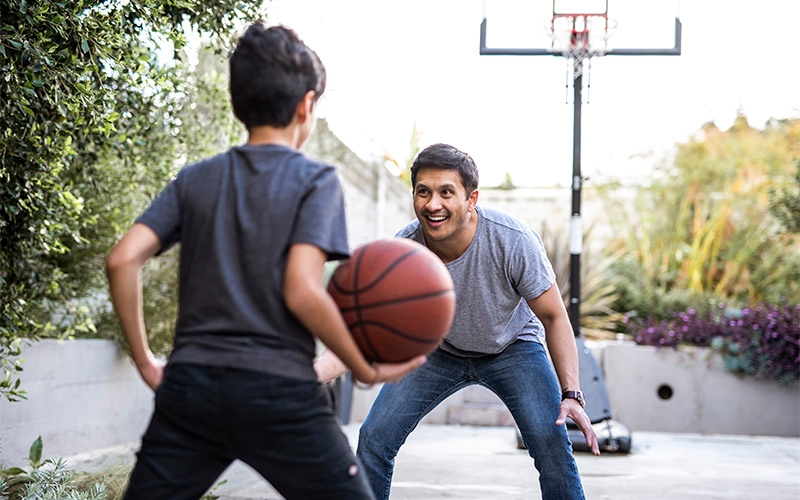Two individuals playing basketball in an outdoor home setting. The foreground shows the back of a person wearing a dark shirt, holding a basketball and preparing to make a move. In the background, another person is standing in a defensive stance.