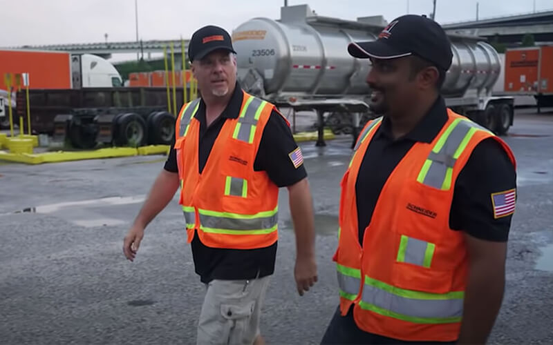 A Driver Mentor and his mentee wearing black shirts, orange safety vests and black baseball hats, walk in a truck yard.