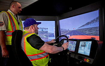 A truck driving student wearing a neon safety vest sits behind the wheel of a training simulator while a driver instructor wearing a tan shirt and neon safety vest watches over his shoulder.