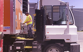 A Yard Jockey wearing jeans, a neon safety vest and black baseball hat hooks up a white terminal trailer to an orange Schneider trailer.