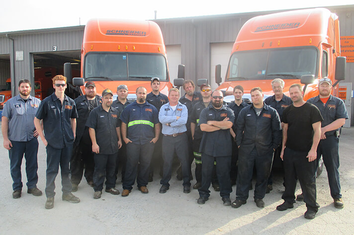 A group of Schneider diesel technicians gathers outside a company shop in front of two company trucks