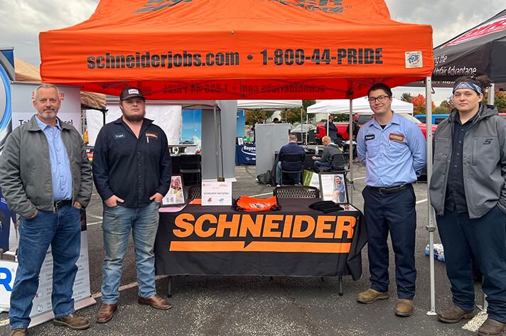 Four Schneider associates, including diesel technicians, stand at a Schneider booth for a hiring event