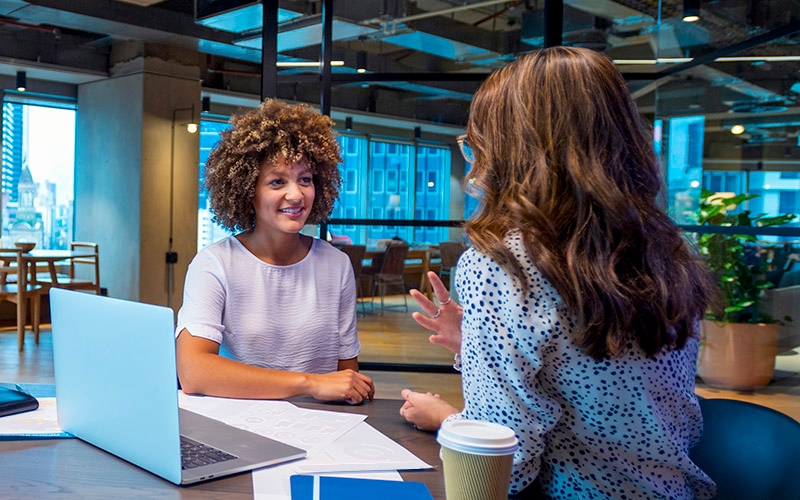 Two women sitting at a table, facing one another and talking in an office setting.