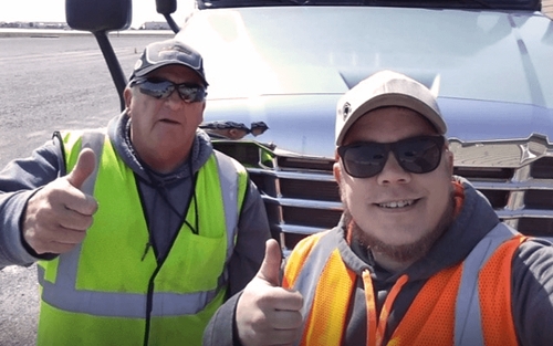 Two male drivers wearing baseball hats, sunglasses, grey hoodies and neon safety vests smile and give a thumbs up while standing in front of a white semi-truck.